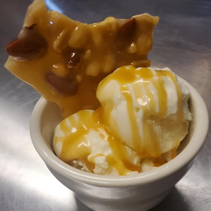Vanilla ice cream with caramel drizzle and a piece of peanut brittle in a white bowl.