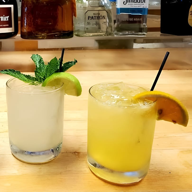 Two Compadres cocktails, one garnished with mint and lime, the other with a lemon slice, on a bar.