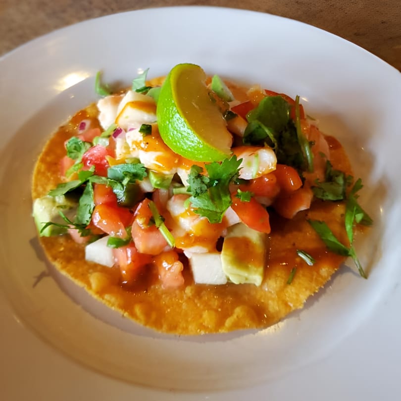 Compadres' shrimp ceviche tostada with avocado, tomato, onion, cilantro, and lime on a white plate.